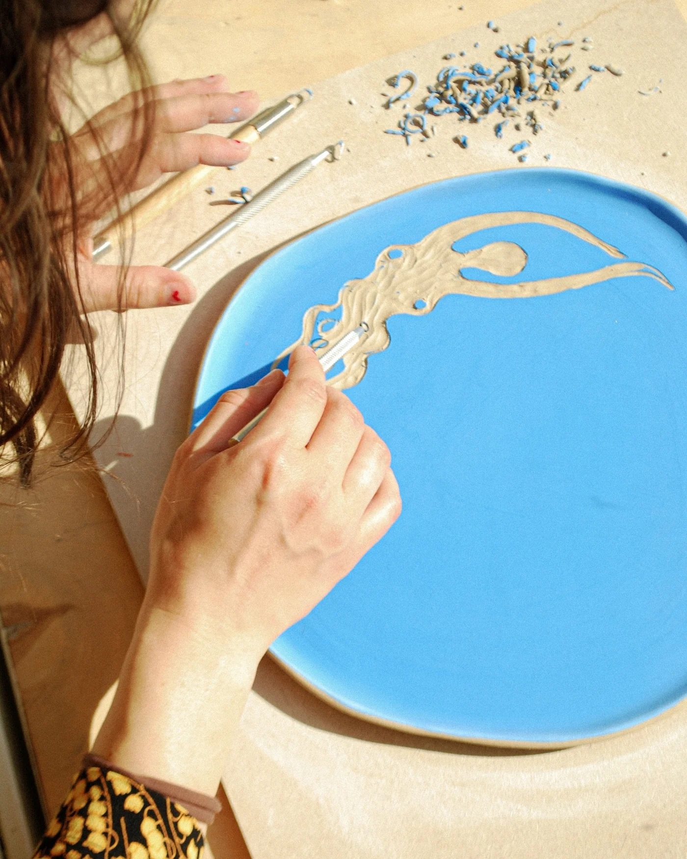 An illustrator carving a body form into a vibrant blue ceramic plate